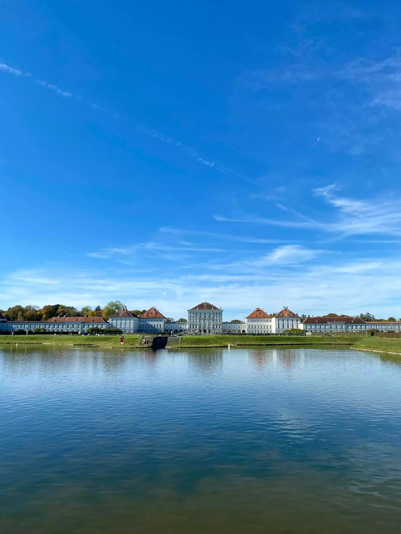 Nymphenburg Palace in Munich, Germany, reflected in the calm waters of its grand canal, with the Baroque façade, manicured gardens, and clear sky creating a symmetrical and tranquil scene.