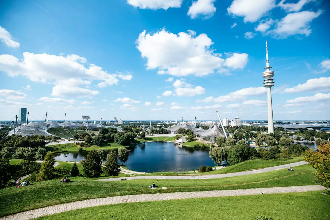 Olympiapark in Munich, Germany, featuring sweeping green lawns, the Olympic Stadium with its iconic tent-like roof structure, and a central lake reflecting the surrounding architecture and trees.
