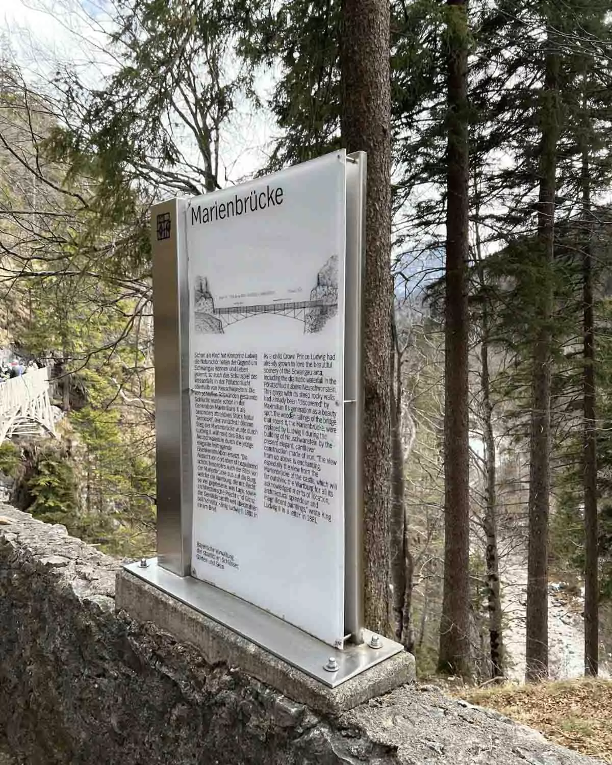 A weathered informational sign detailing the history of Marienbrücke, a famous pedestrian bridge in Germany. The sign is mounted on a wooden post with natural surroundings visible in the background.