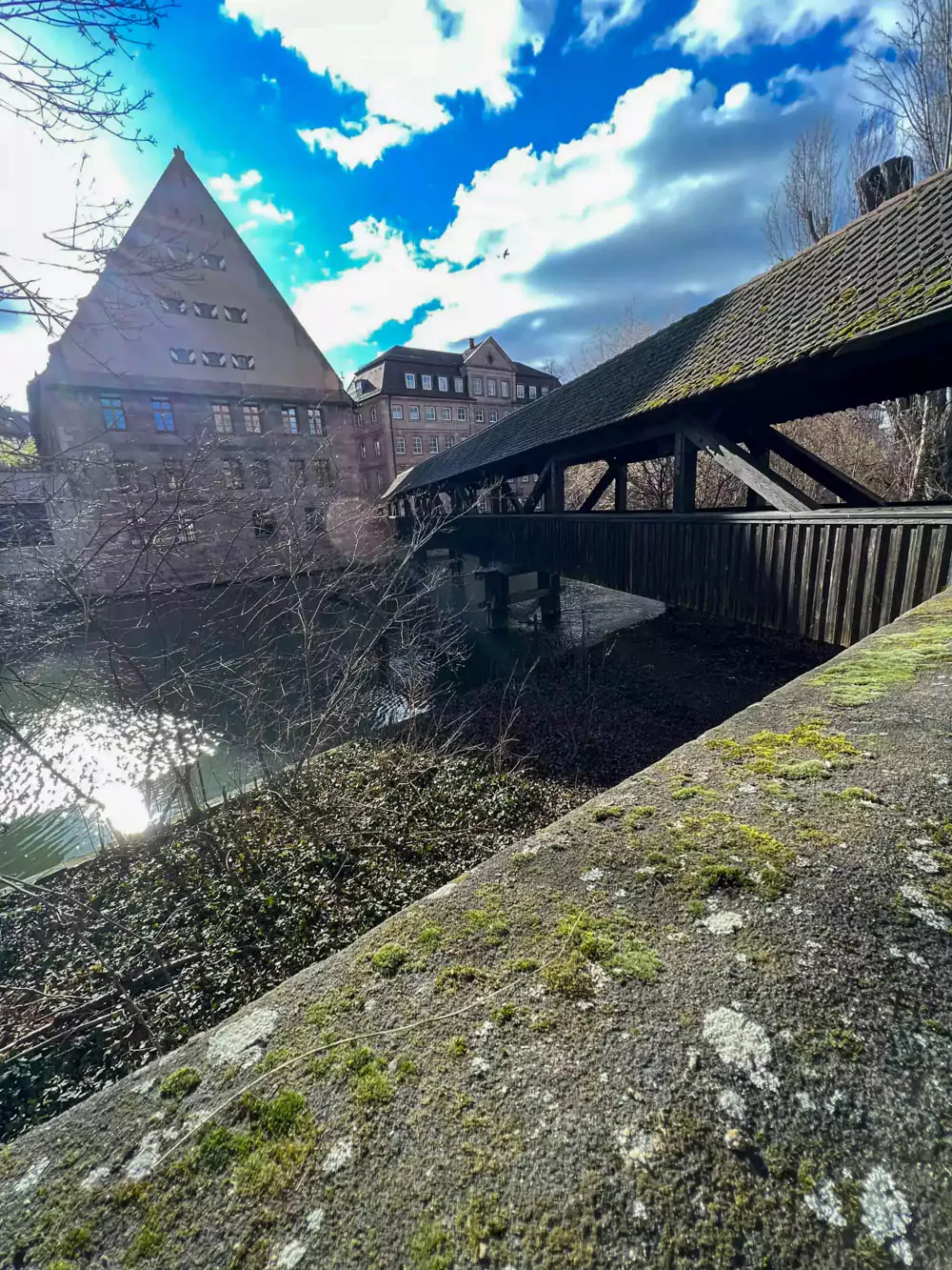 Hangman’s Bridge (Henkersteg) in Nuremberg, Germany, a covered wooden bridge crossing the river Pegnitz, with timber framing and stone foundations, historically linked to the city’s executioner’s residence and offering a view of medieval architecture along the water.