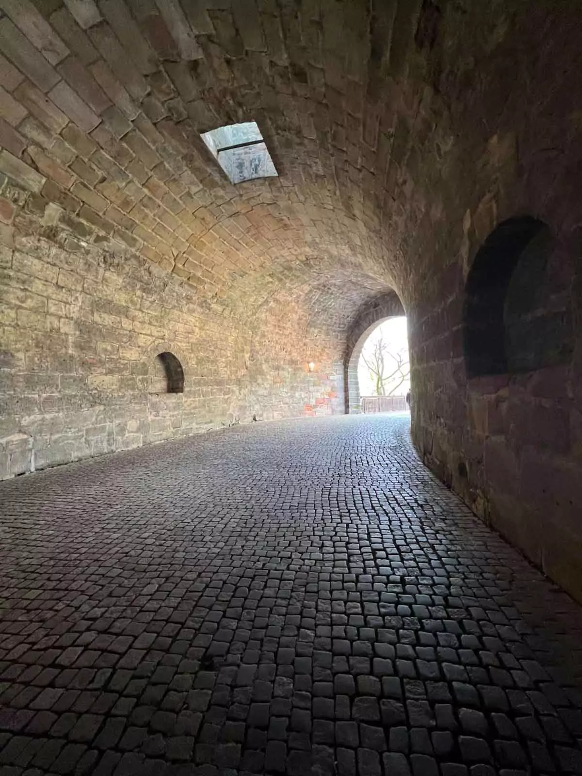A narrow stone tunnel passes through the medieval city walls of Nuremberg, with rough-hewn stones forming the archway and walls. The historic passageway leads into a cobblestone street lined with traditional buildings.