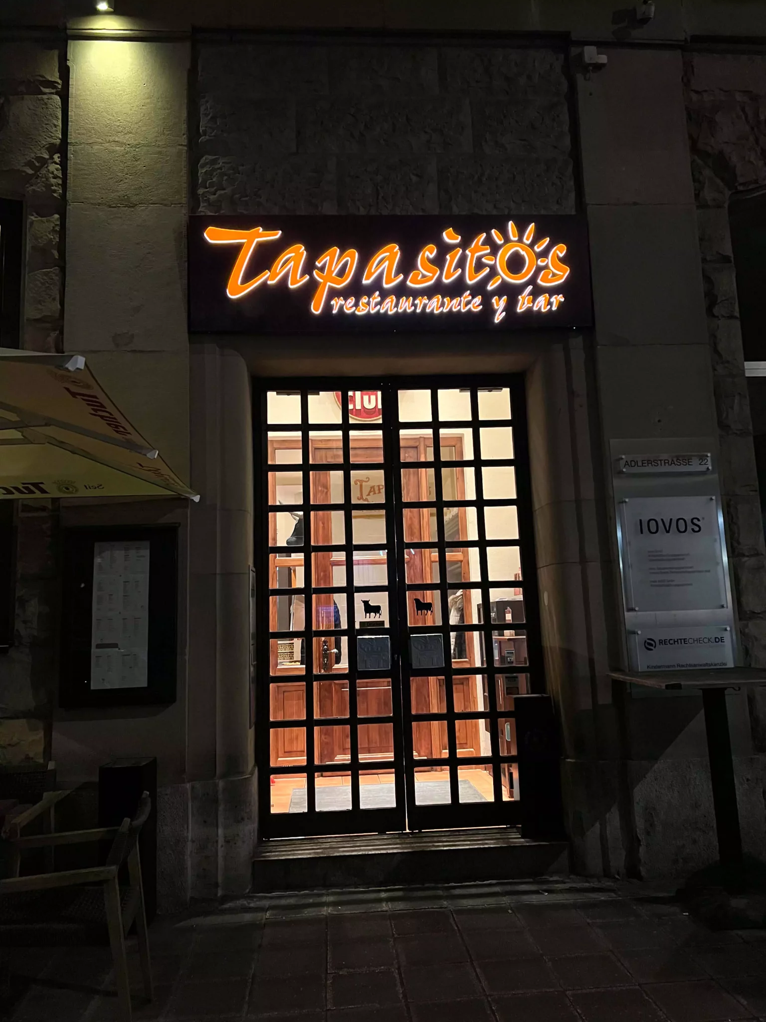 The entrance to Tapasitos in Nuremberg, Germany, illuminated at night with warm, inviting lighting. The modern glass facade and subtle signage create a cozy yet stylish vibe for this Spanish-inspired eatery.