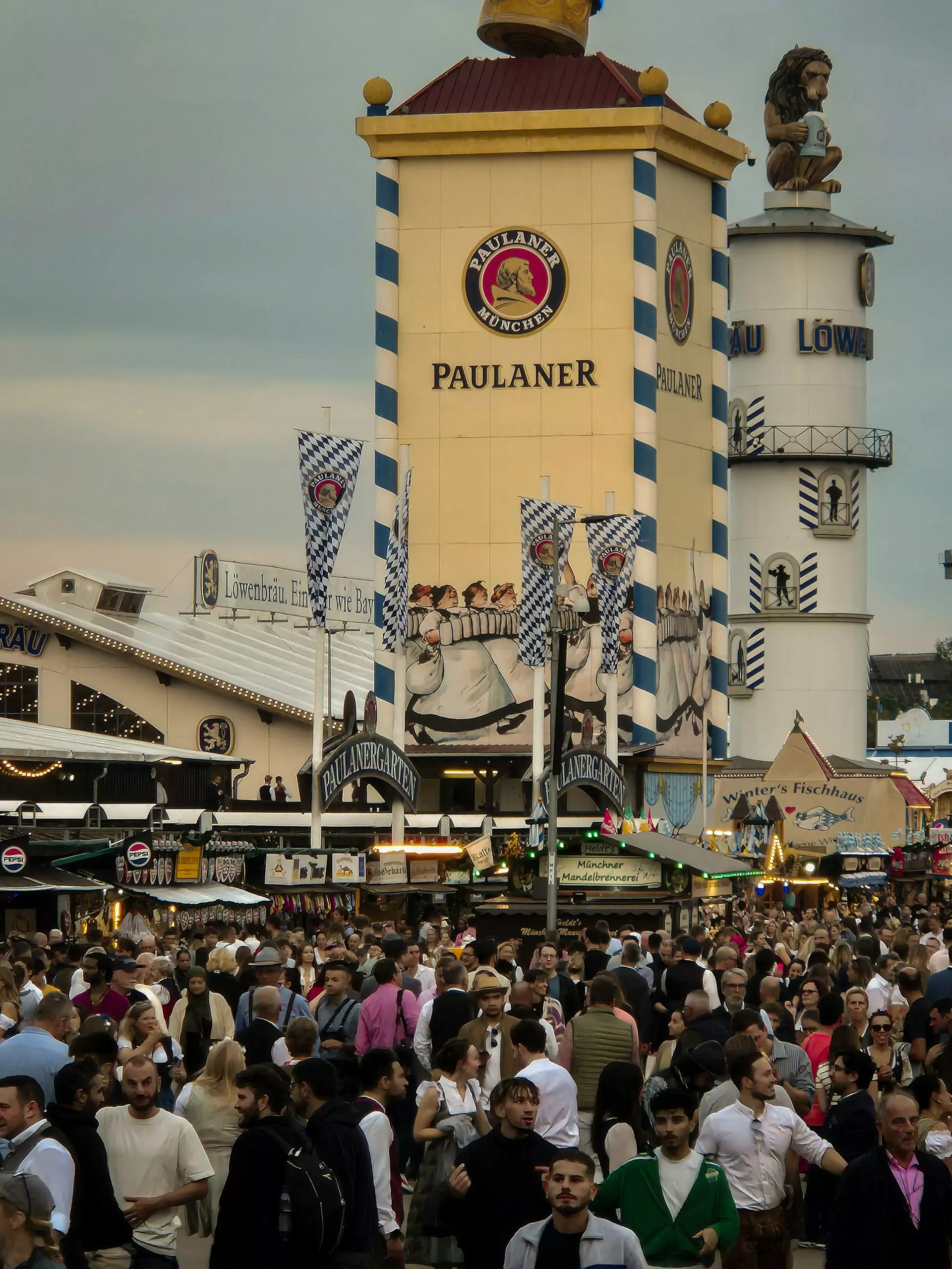 Crowd at Oktoberfest in Munich, Germany, gathered near the iconic Paulaner beer tower, with people in traditional Bavarian attire enjoying the celebration under bright skies and festive decorations.