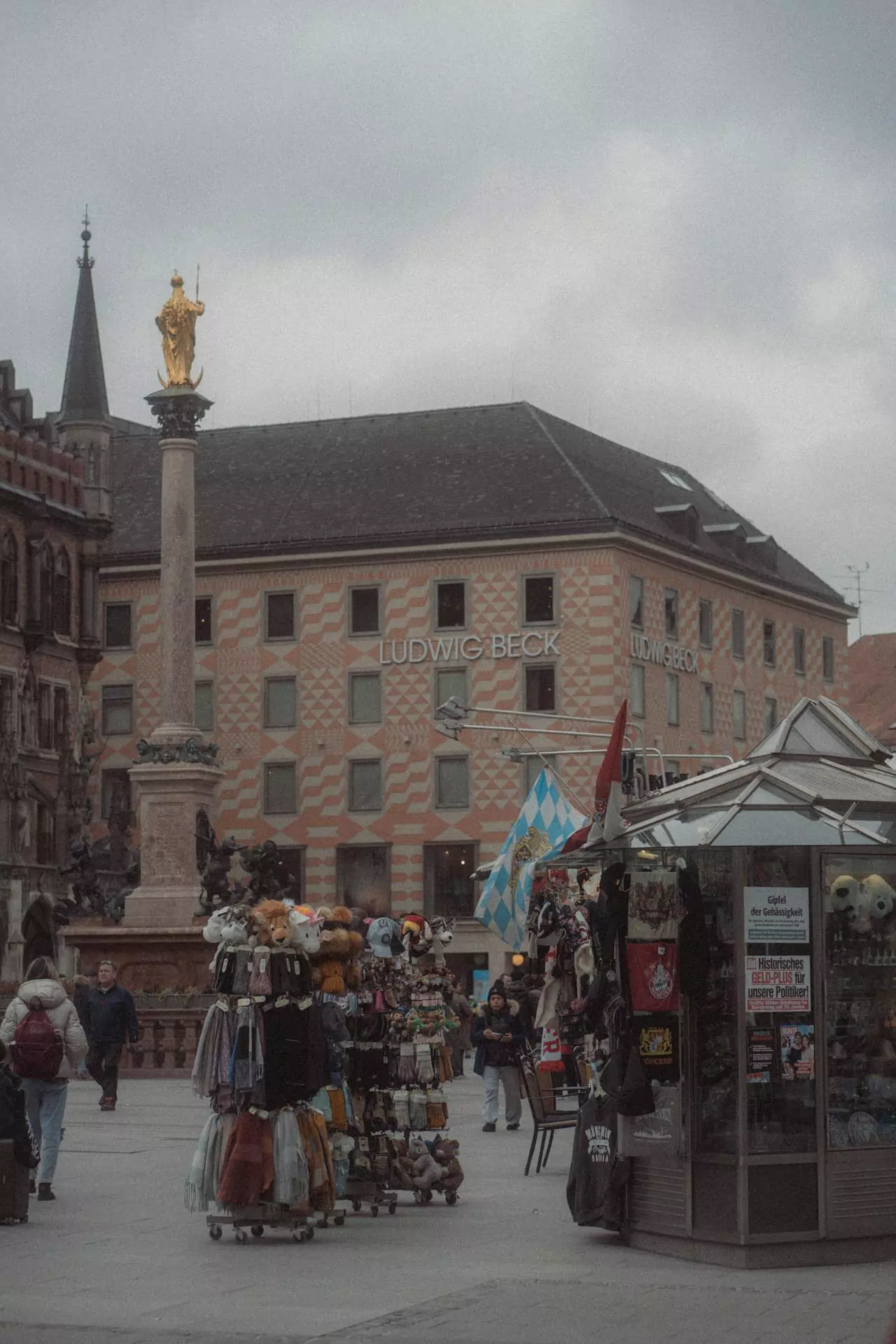 People shopping in Marienplatz, Munich, Germany, walking past stores and stalls in the historic city square, with the New Town Hall and Mariensäule providing a scenic backdrop to the bustling atmosphere.