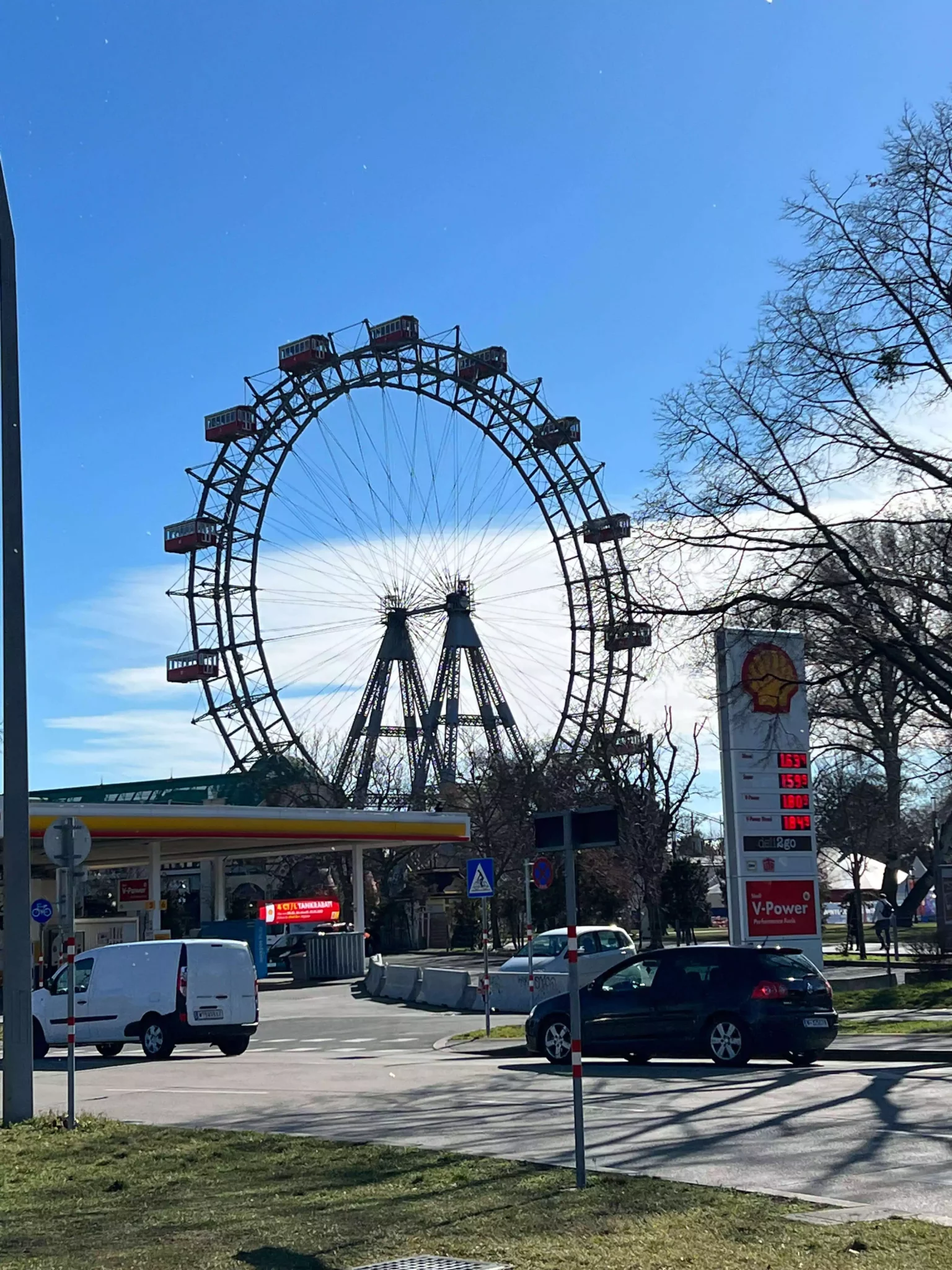 The giant Ferris wheel at Prater amusement park in Vienna, Austria, rises in the background behind a nearby gas station. The contrast between the historic fairground ride and the modern fuel stop creates a quirky urban juxtaposition.