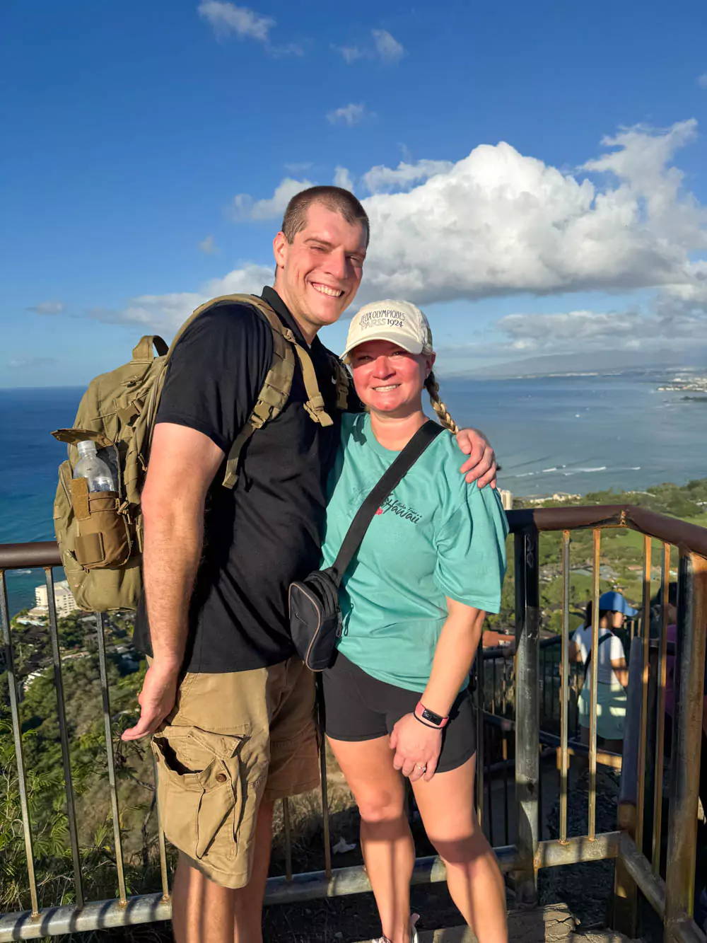 Katie and Zack at the summit of Diamond Head in Oʻahu, Hawaii, in December, smiling with panoramic views of Waikīkī, the coastline, and blue Pacific Ocean behind them.