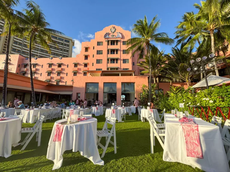 Exterior of The Royal Hawaiian hotel in Waikīkī, known as the “Pink Palace of the Pacific,” with its signature pink stucco facade framed by palm trees and tropical gardens.