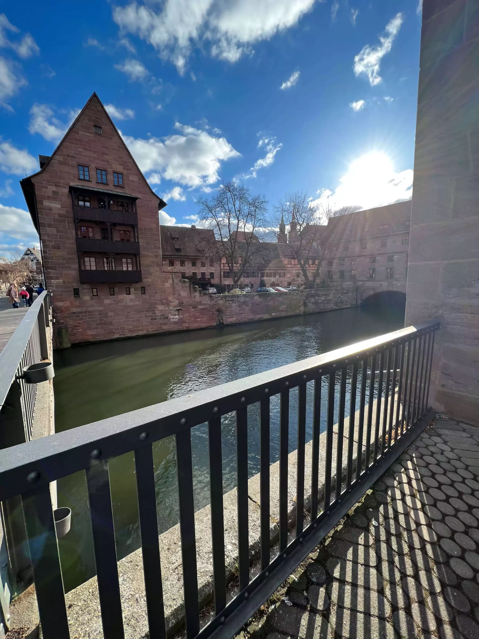 Heilig-Geist-Spital in Nuremberg, Germany, viewed from the east, with its arched stone structure spanning the Pegnitz River. The medieval building’s timber-framed upper floors and red-tiled roof reflect beautifully in the calm water below.
