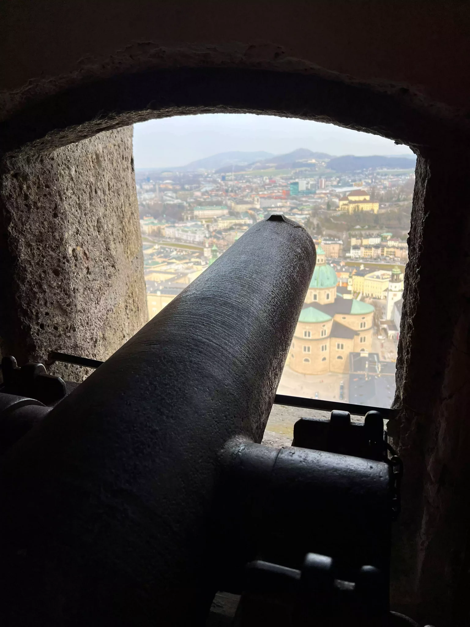 Historic cannon positioned inside an arched stone opening at Hohensalzburg Fortress, overlooking the rooftops and church spires of Old Town Salzburg, Austria.