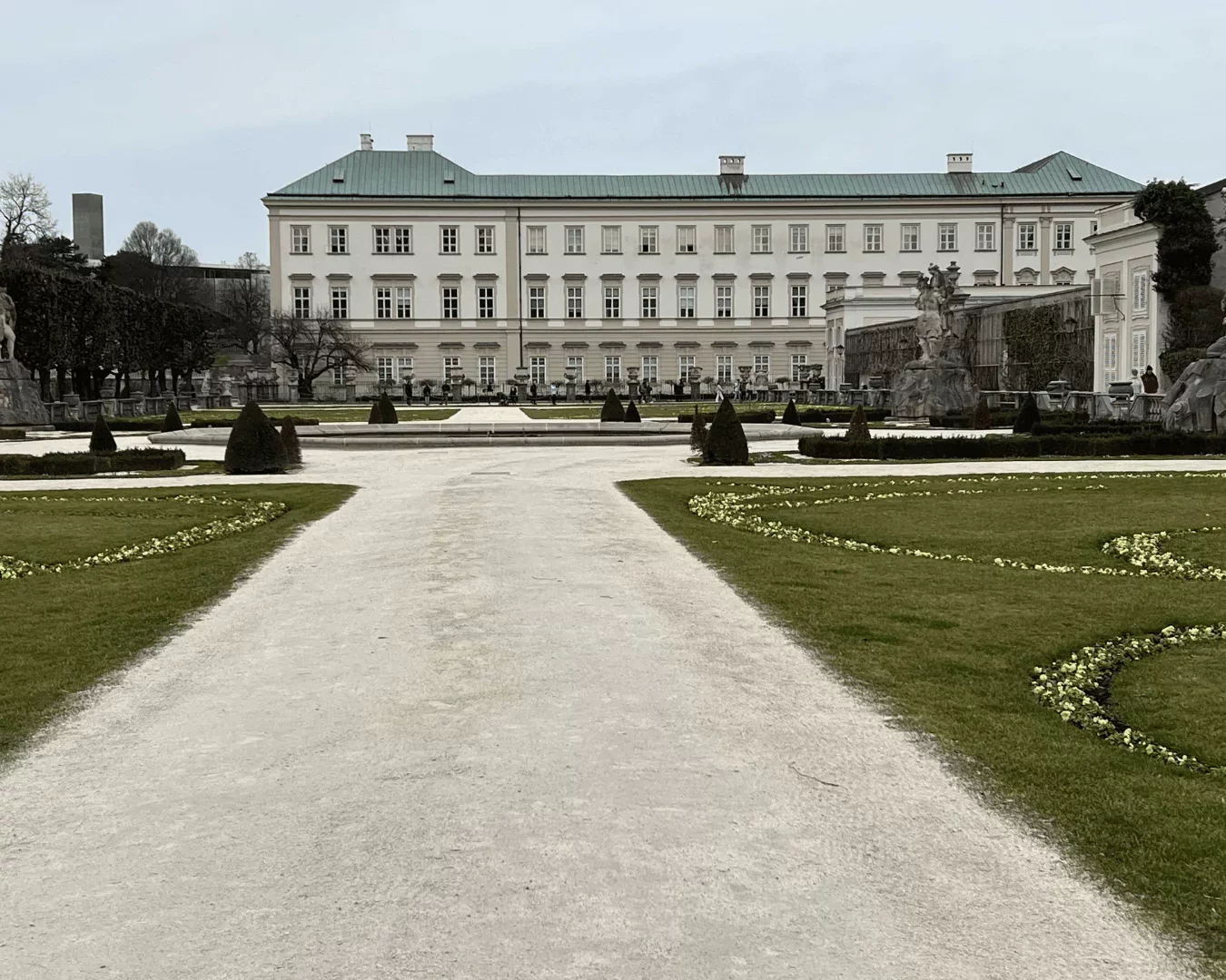 The historic Mirabell Palace in Salzburg, Austria, stands elegantly behind its beautifully landscaped gardens, now dusted with winter frost. Bare trees and trimmed hedges frame the quiet, symmetrical paths, offering a serene view of the palace in the colder season.