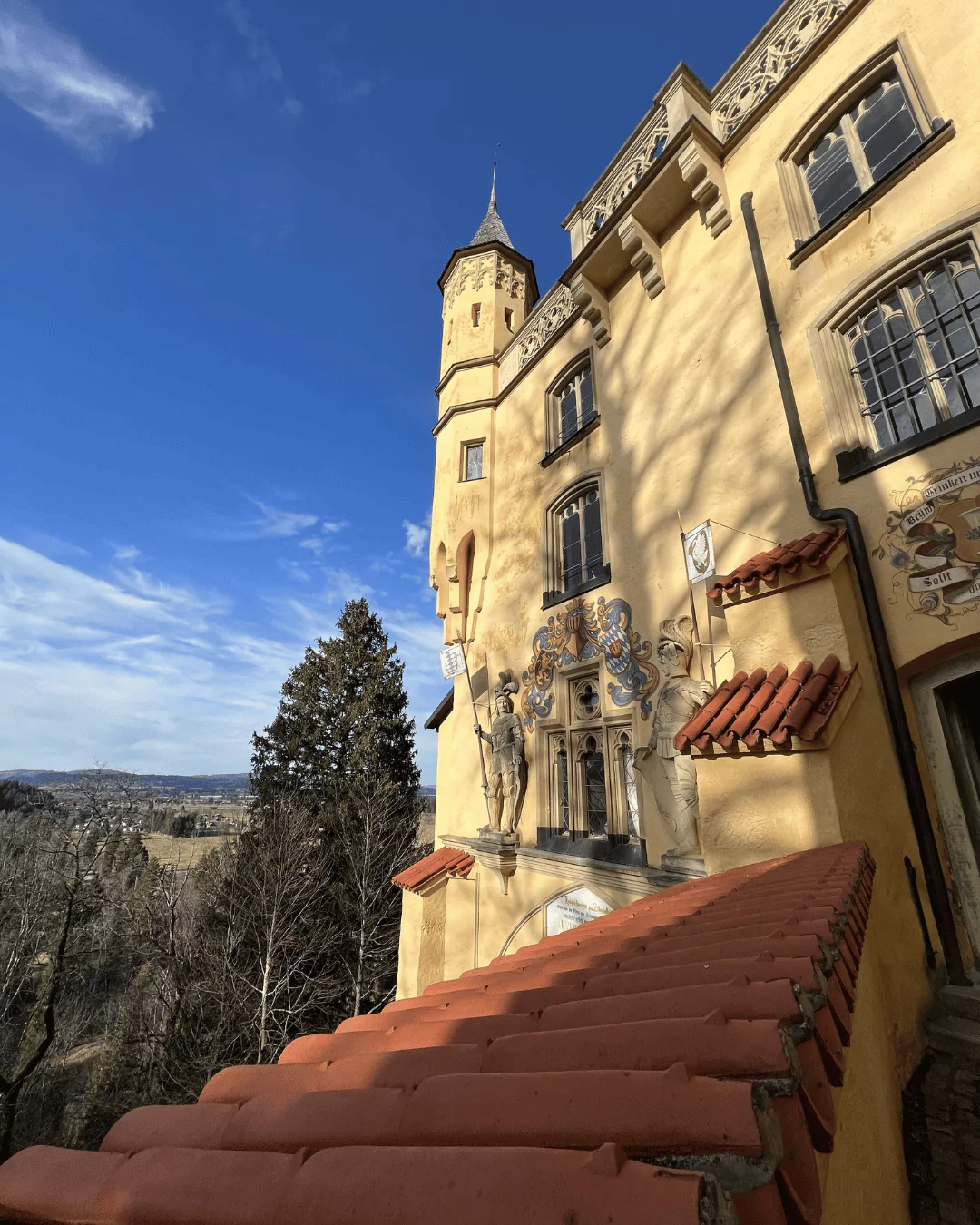 Exterior of Hohenschwangau Castle in Bavaria, Germany, showcasing its vibrant yellow facade adorned with statues, ornate window frames, and decorative medieval-style murals and embellishments.
