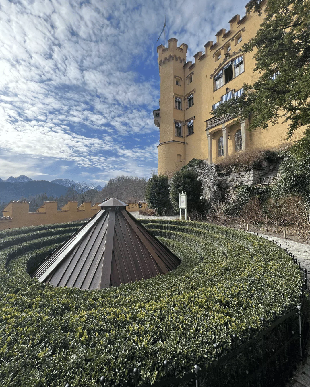 Hohenschwangau Castle gardens in Bavaria glowing under a bright blue sky, with manicured greenery, stone pathways, and views of the yellow castle walls.