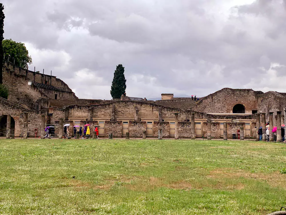 Amphitheatre of Pompeii, Italy, featuring a large elliptical arena with stone seating tiers and arched entryways, one of the oldest surviving Roman amphitheaters, set against a backdrop of cloudy skies.