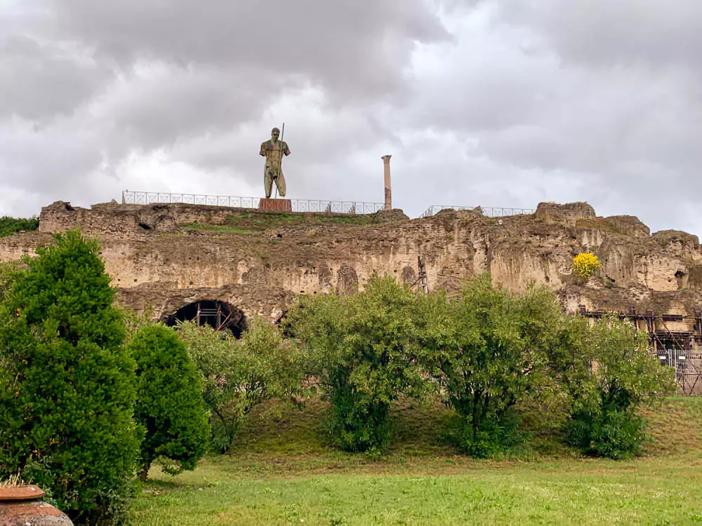 Daedalus statue by Igor Mitoraj in the Archaeological Park of Pompeii, Italy, set against ancient ruins on a cloudy day, with the monumental bronze figure standing in striking contrast to the weathered stone surroundings.