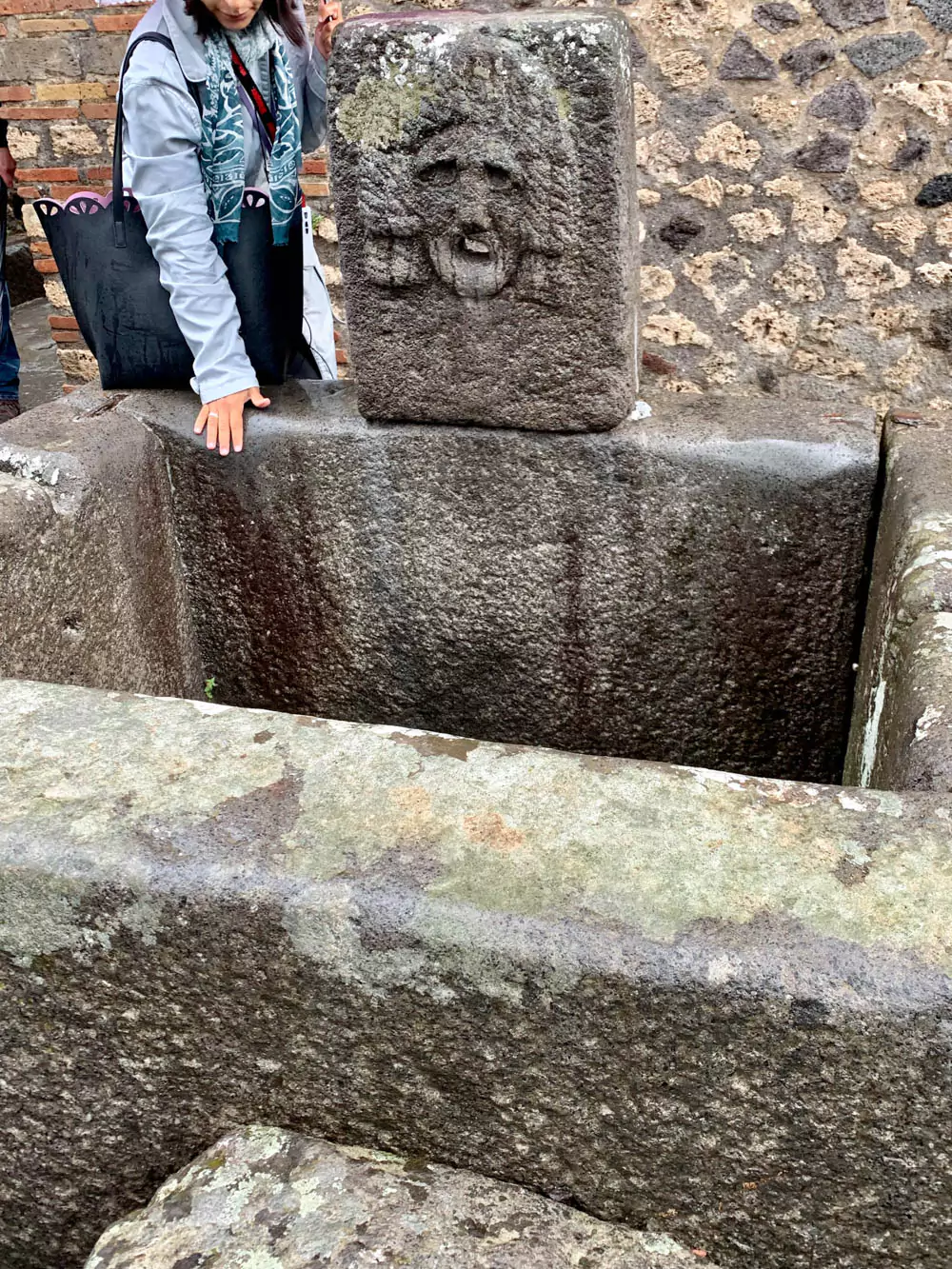 Ancient fountain in Pompeii featuring a carved relief of a tragic theatrical mask, with weathered stone details and a spout once used to pour water, reflecting Roman art and performance culture.