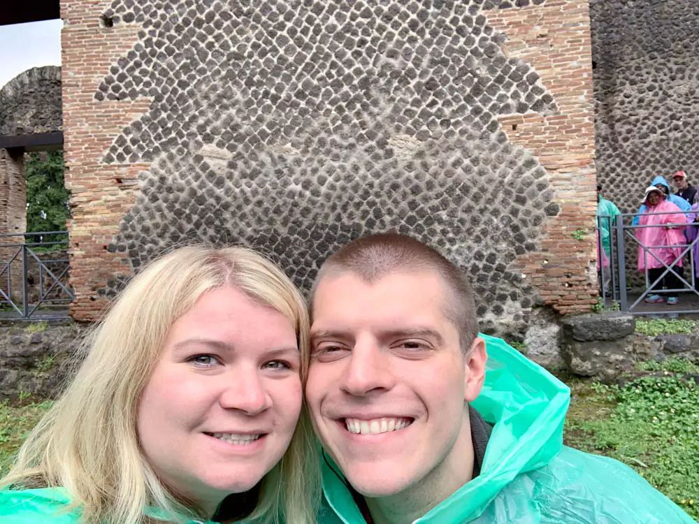 Selfie of Katie and Zack wearing green ponchos at the Archaeological Park of Pompeii in Italy, smiling with ancient ruins and stone pathways visible in the background on a rainy day.