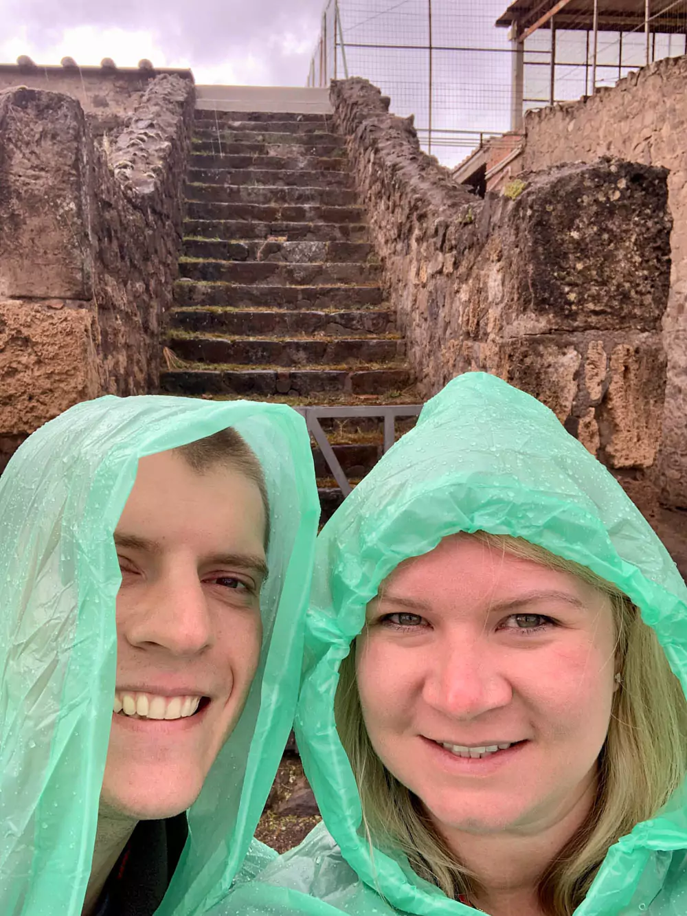 Katie and Zack standing on an ancient stone staircase in the Archaeological Park of Pompeii, Italy, surrounded by weathered ruins and historic Roman architecture on a cloudy day.