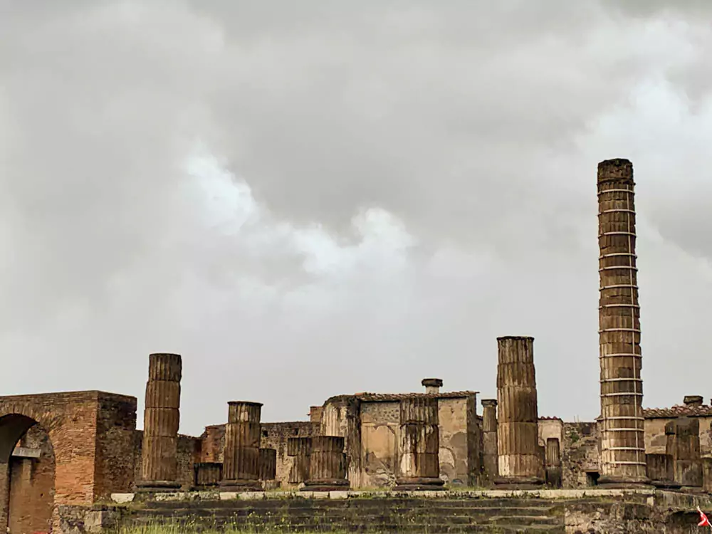 Temple of Jupiter in the Archaeological Park of Pompeii, Italy, on a cloudy day, with ancient stone columns and ruins standing against an overcast sky, while Mount Vesuvius is obscured by clouds in the background.