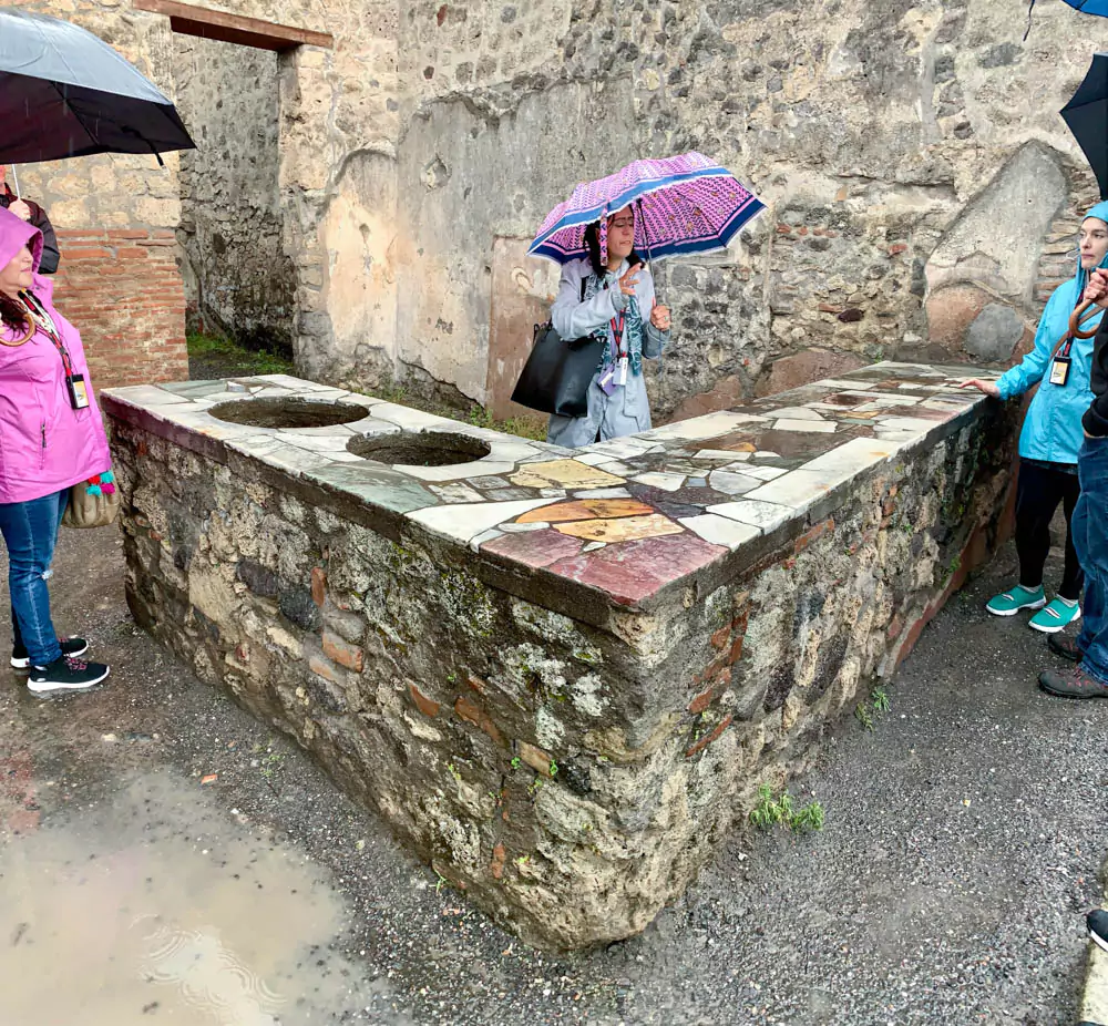 Thermopolium or Caupona of Primus in Pompeii, Italy, showing a well-preserved Roman street food counter with embedded dolia (storage jars), colorful frescoes, and stone countertops used to serve hot food and drinks.