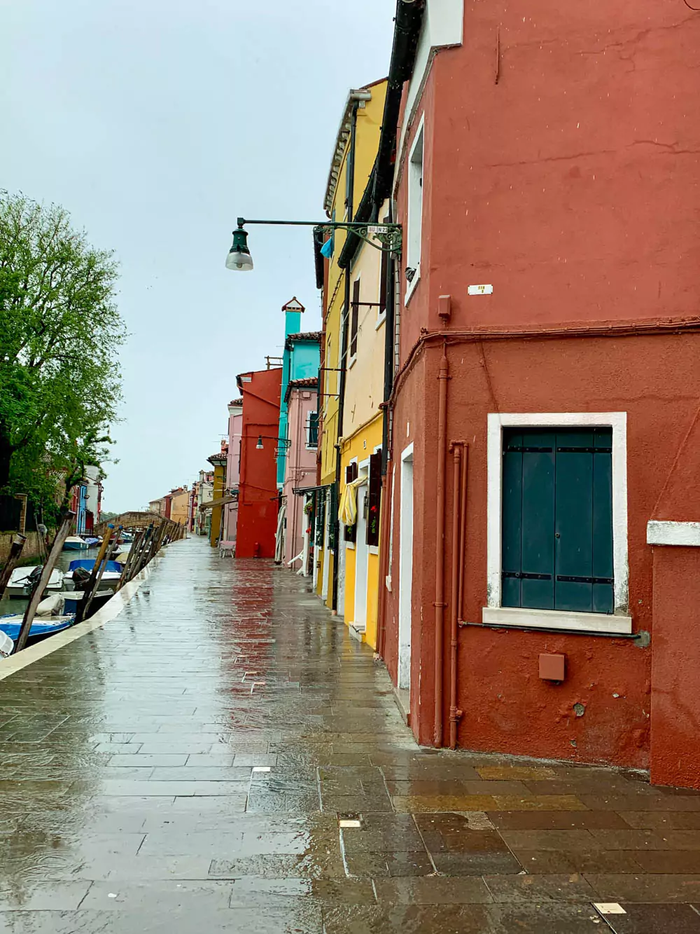 Wet sidewalk in Burano, Italy, reflecting the brightly colored facades of houses lining the canal, with moored boats and soft rain adding to the charm of the island village.