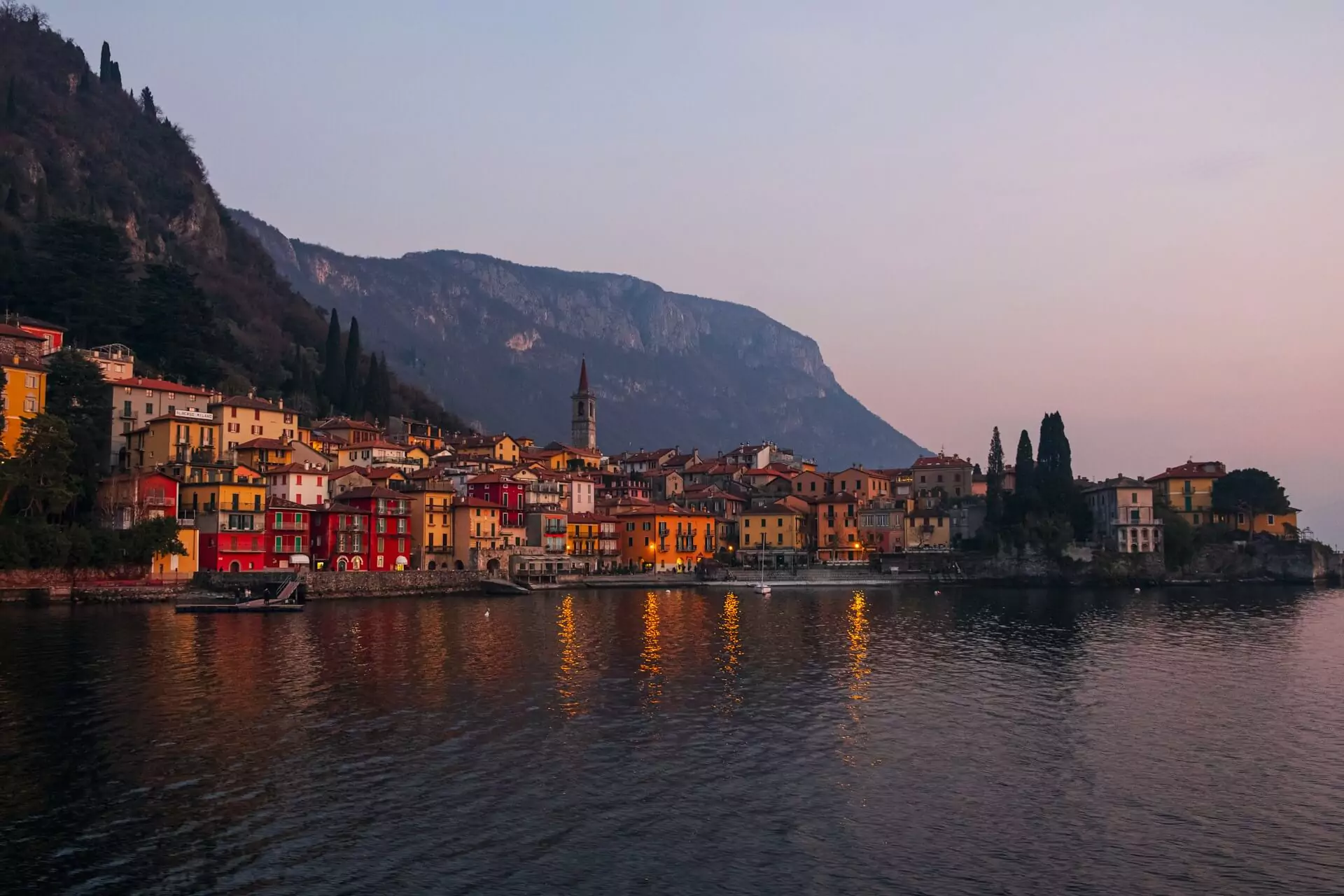 Lake Como at dusk, with the water reflecting soft shades of pink and blue from the fading sky, silhouetted mountains in the background, and lights beginning to twinkle in the lakeside villages