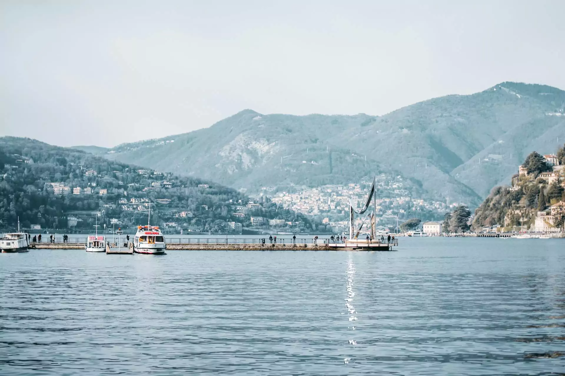 Boats floating on the clear blue waters of Lake Como in Italy on a sunny day, with scenic hillsides and colorful lakeside buildings in the background under a clear sky.