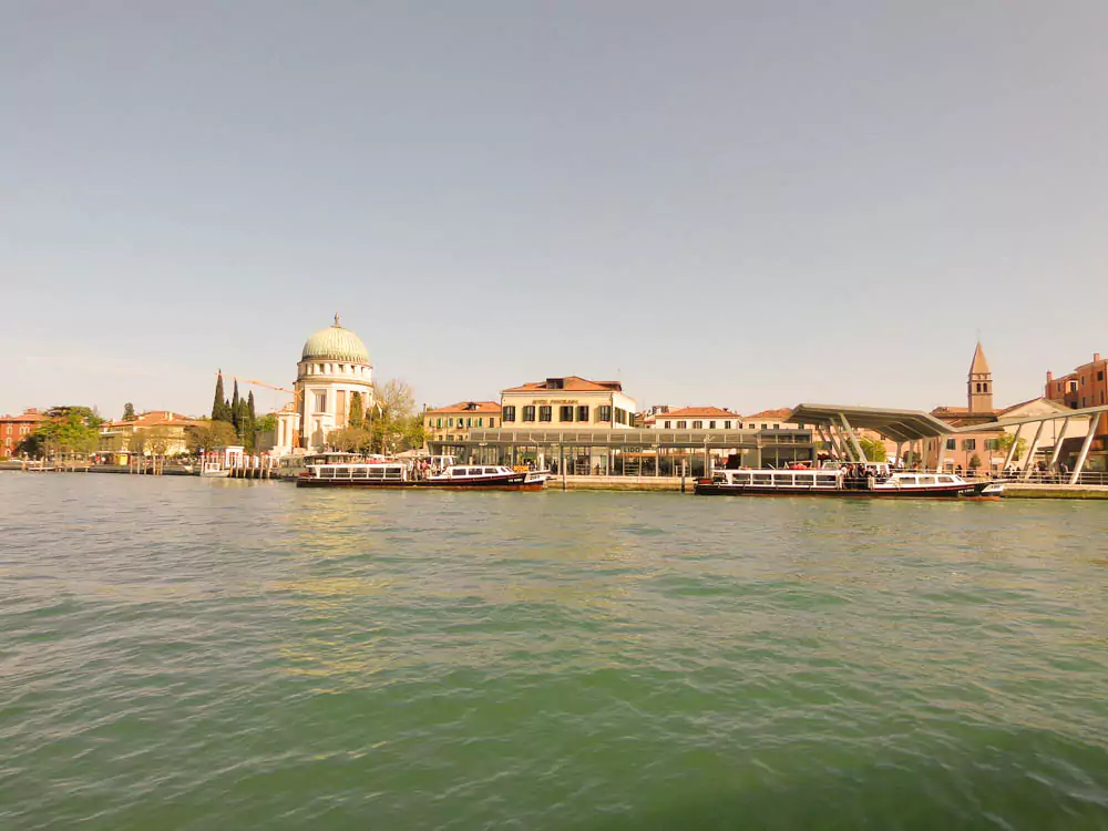 A view of Lido di Venezia from the lagoon, featuring a domed waterfront structure surrounded by small boats and gentle waves, with the island’s coastline stretching into the distance