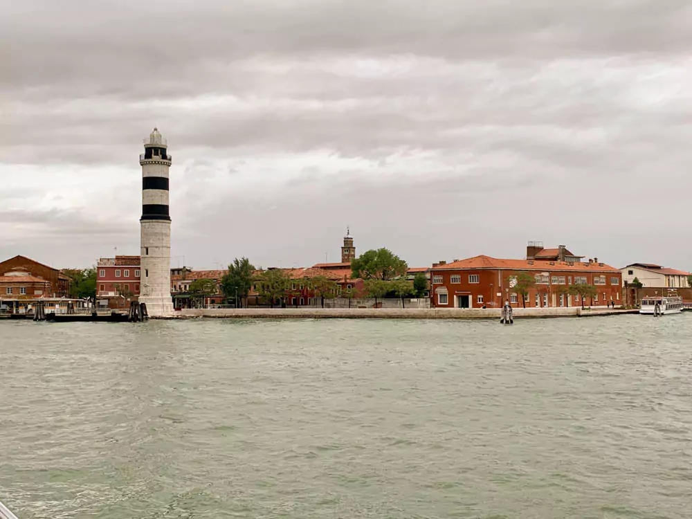 View of Murano, Italy, from the Venetian Lagoon, featuring colorful waterfront buildings, small docks, and a peaceful stretch of water under a bright, open sky.