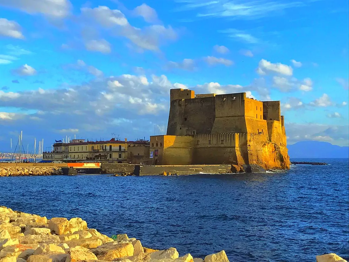 Castel dell'Ovo in Naples, Italy, perched on a rocky islet and surrounded by deep blue waters of the Gulf of Naples, with stone walls rising above the sea under a clear sky.