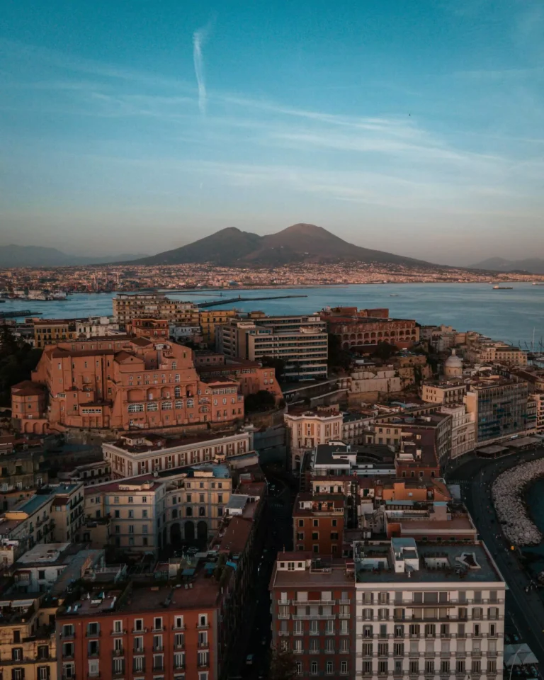 Panoramic view of Naples, Italy, with a dense array of colorful buildings climbing the hillside, Mount Vesuvius in the background, and the Bay of Naples stretching into the distance under a sunny sky.
