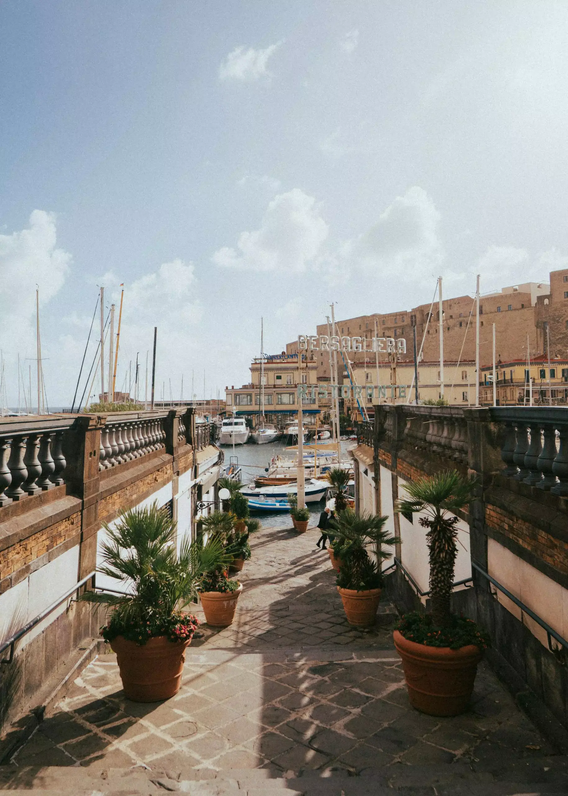 View of a marina in Naples, Italy, with boats docked along calm blue waters, backed by colorful waterfront buildings and distant hills under a clear, sunny sky.