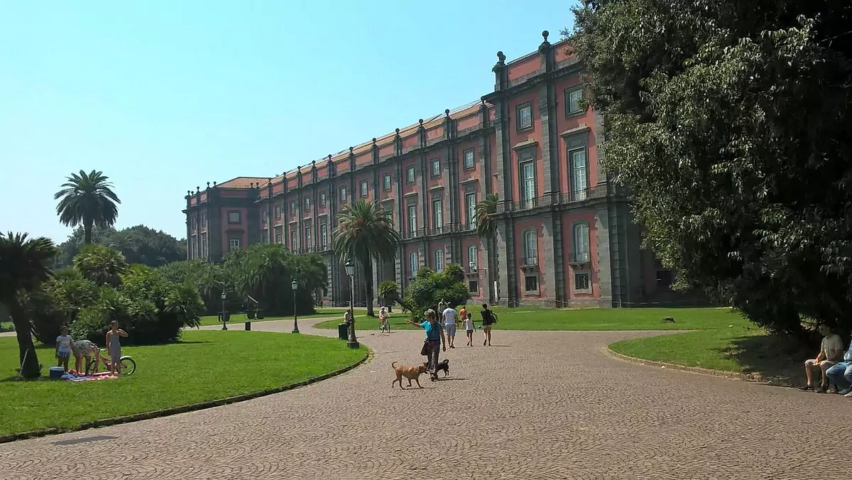 Exterior of the Museum of Capodimonte in Naples, Italy, housed in a grand Bourbon-era palace with a red and gray stone façade, surrounded by manicured gardens and known for its extensive art collection.