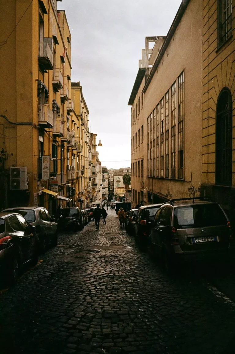 Old buildings lining a narrow city street in Naples, Italy, with colorful facades, laundry hanging from balconies, and scooters parked along the cobblestone path, capturing the charm of everyday Neapolitan life.