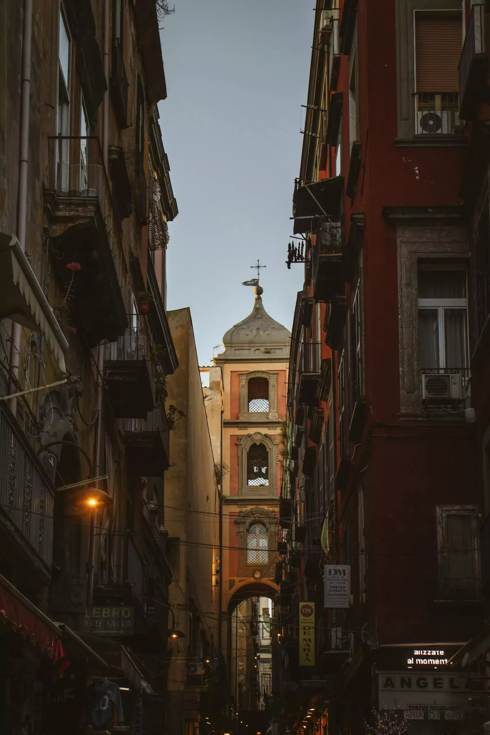 San Gregorio Armeno Church at dusk in Naples, Italy, with its Baroque façade softly illuminated against the fading light, as the surrounding street begins to glow with warm evening tones.