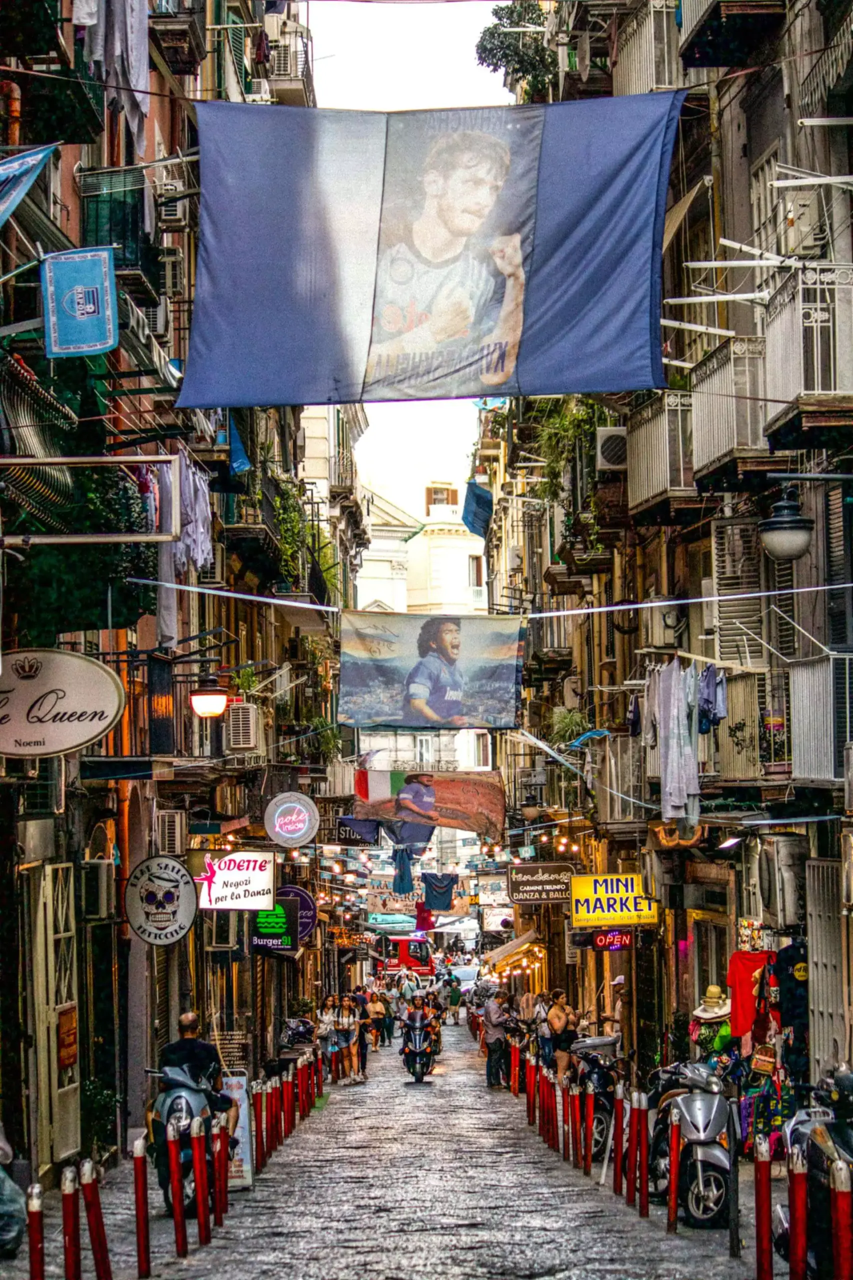 Charming street in old town Naples, Italy, lined with aged stone buildings, wrought-iron balconies, and bustling with local life, scooters, and hanging laundry above the narrow walkway.