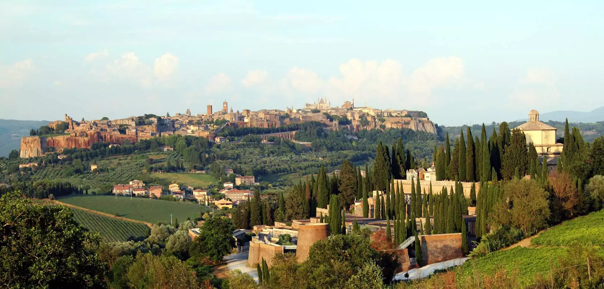 View of the hilltop town of Orvieto, Italy, in summer, with its medieval stone buildings and cathedral rising above lush green hills and terraced countryside under a bright blue sky.