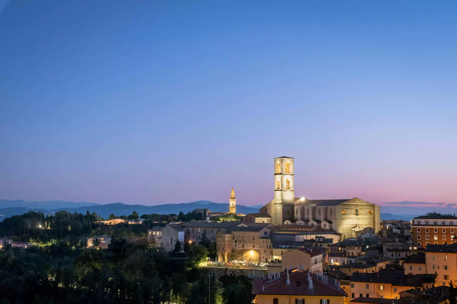 Evening panorama of Perugia, Italy, with the Convent of San Domenico prominently visible among the city’s terracotta rooftops and historic buildings, all bathed in warm, golden-hour light under a soft sky.