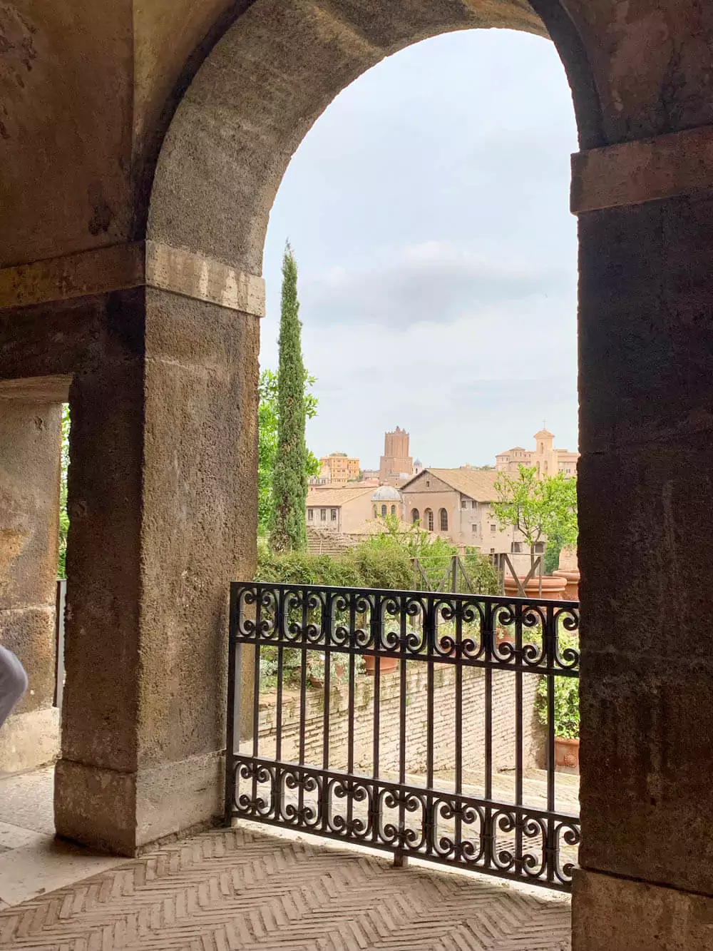 Glimpse of the Roman Forum through an arched open window, framing ancient ruins, stone columns, and historic pathways under a bright, clear sky, offering a picturesque view of Rome’s past.