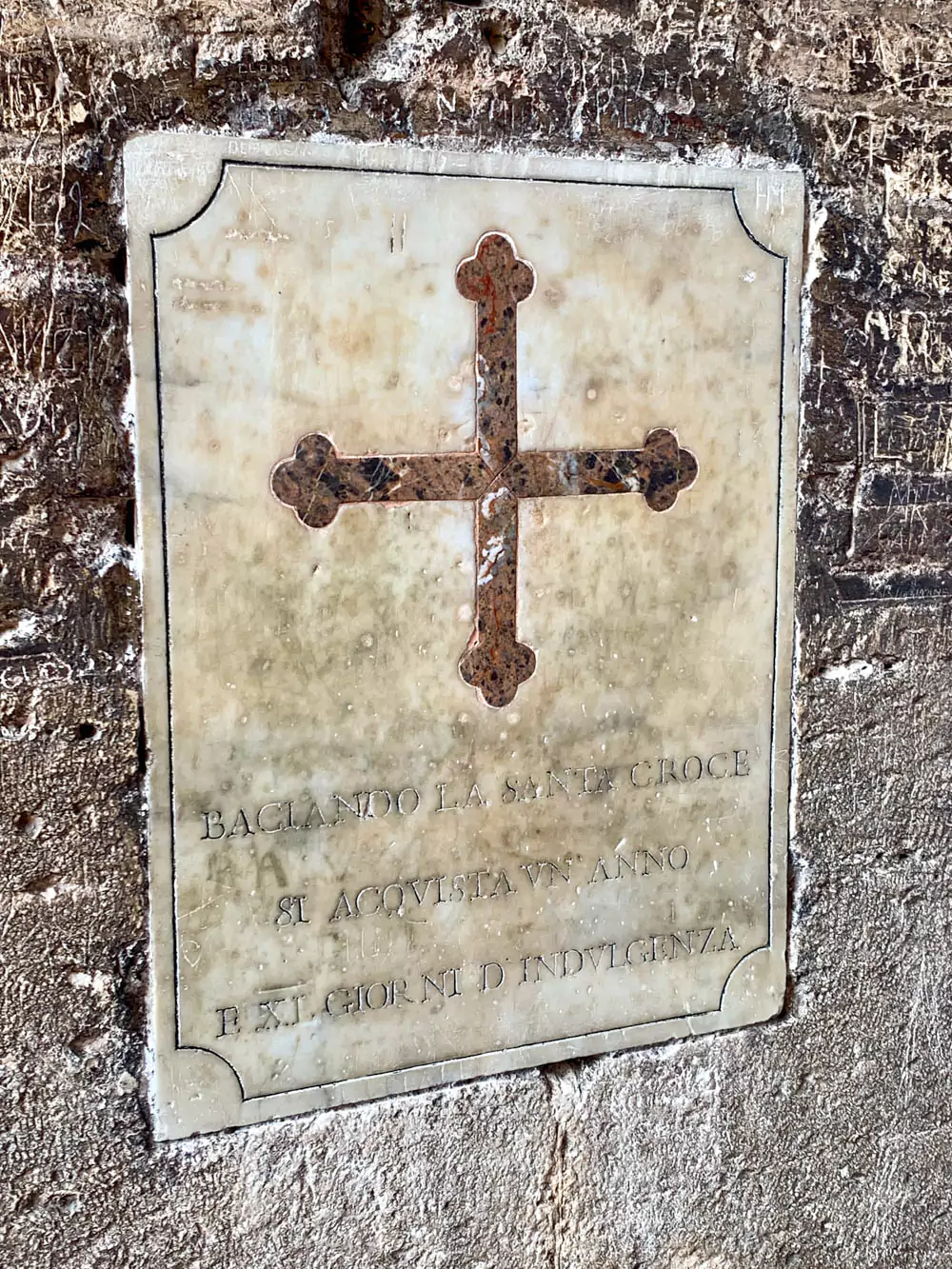 Ancient Italian sign mounted on a wall inside the Colosseum in Rome, reading “Kissing the Cross you will receive one year and eleven days of indulgence,” reflecting a blend of Christian devotion and historical tradition within the ancient amphitheater.