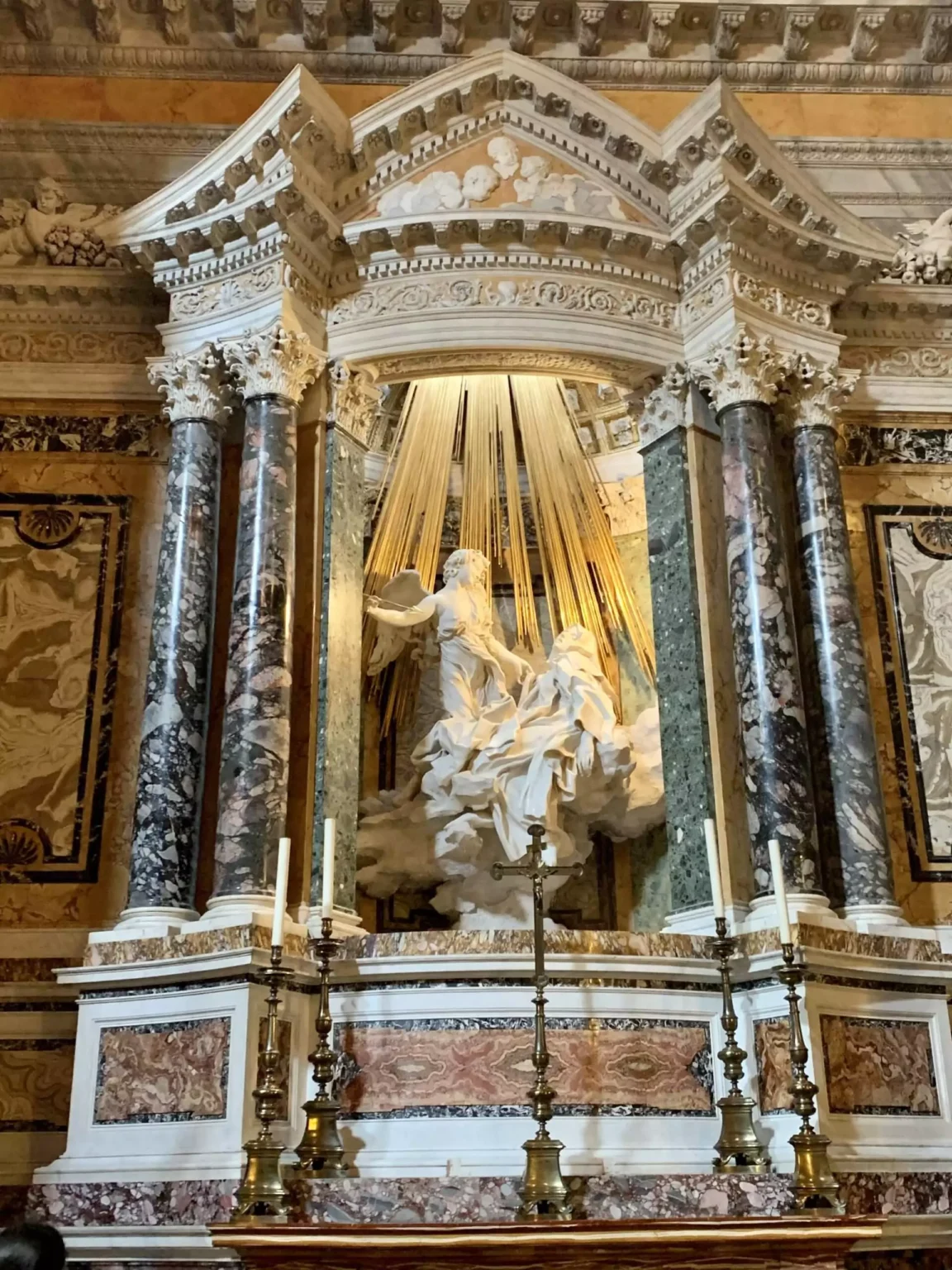 Bernini’s Ecstasy of Saint Teresa in Santa Maria della Vittoria, Rome, depicting the saint in a moment of divine rapture with an angel poised beside her. The white marble sculpture is dramatically lit and framed by golden rays, showcasing Baroque artistry and spiritual intensity.