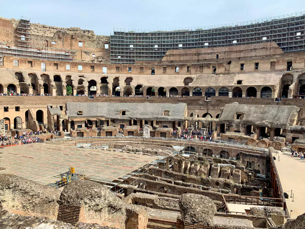 View of the Colosseum's arena floor, showing the reconstructed wooden platform and underground passages, surrounded by ancient stone arches and seating tiers.