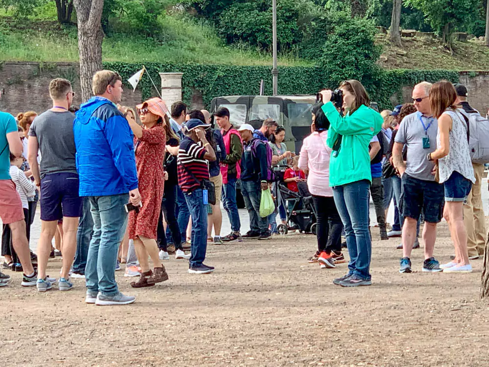 Crowds of tourists queueing outside the Colosseum in Rome, gathering near the ancient stone arches under a sunny sky, waiting to enter the historic Roman amphitheater.