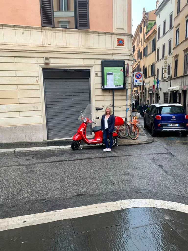 Katie standing next to a bright red Vespa scooter on a cobblestone street in Rome, with historic buildings and a charming urban backdrop capturing the classic Italian vibe.
