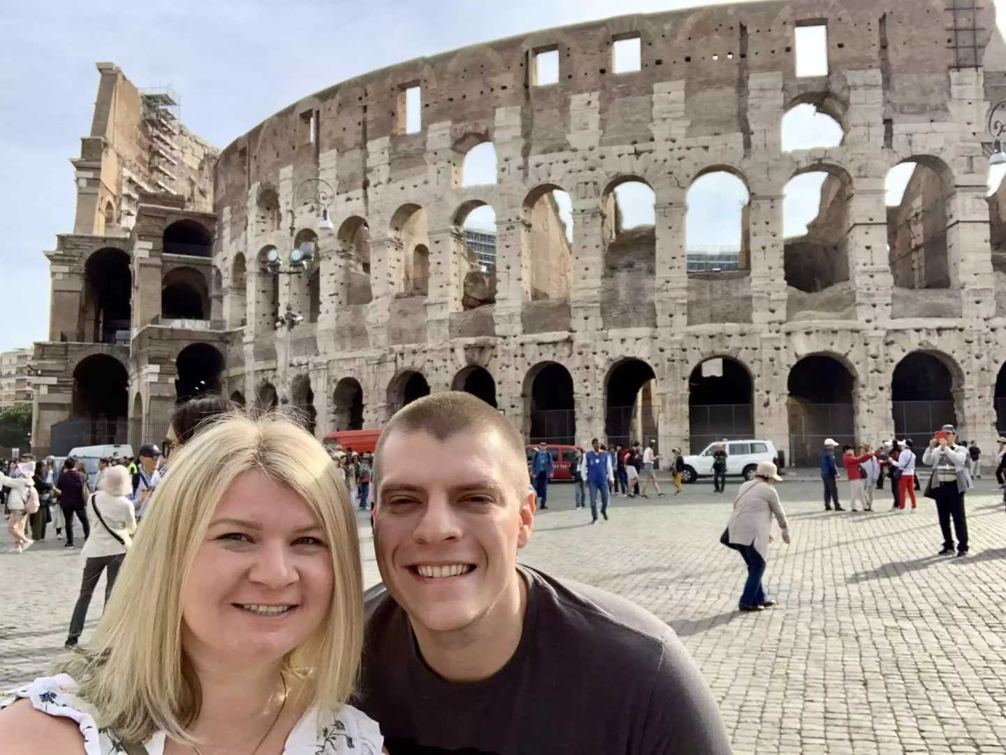 Katie and Zack posing together in front of the Colosseum in Rome, smiling with the iconic ancient stone arches towering behind them under a bright sky. The historic amphitheater creates a dramatic and timeless backdrop for their joyful moment.