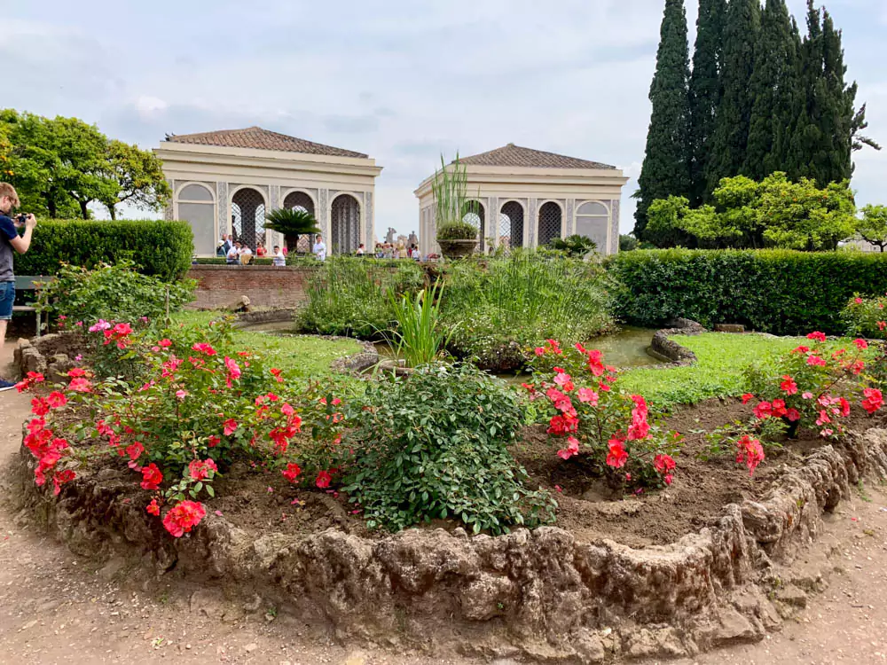 Two charming gazebos set within the gardens of Palatine Hill in Rome, surrounded by manicured greenery, tall trees, and ancient ruins, offering peaceful spots to rest amid the historic landscape.