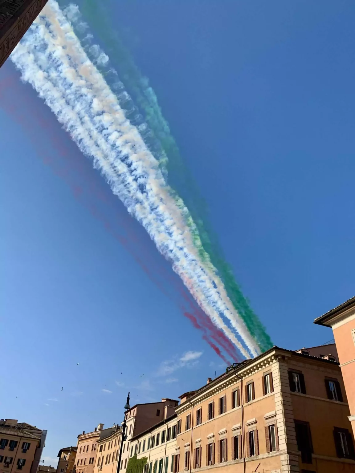 Jets flying in formation over Piazza Navona in Rome, leaving vibrant trails of green, white, and red smoke representing the colors of the Italian flag. The patriotic display contrasts against the blue sky and historic buildings below.