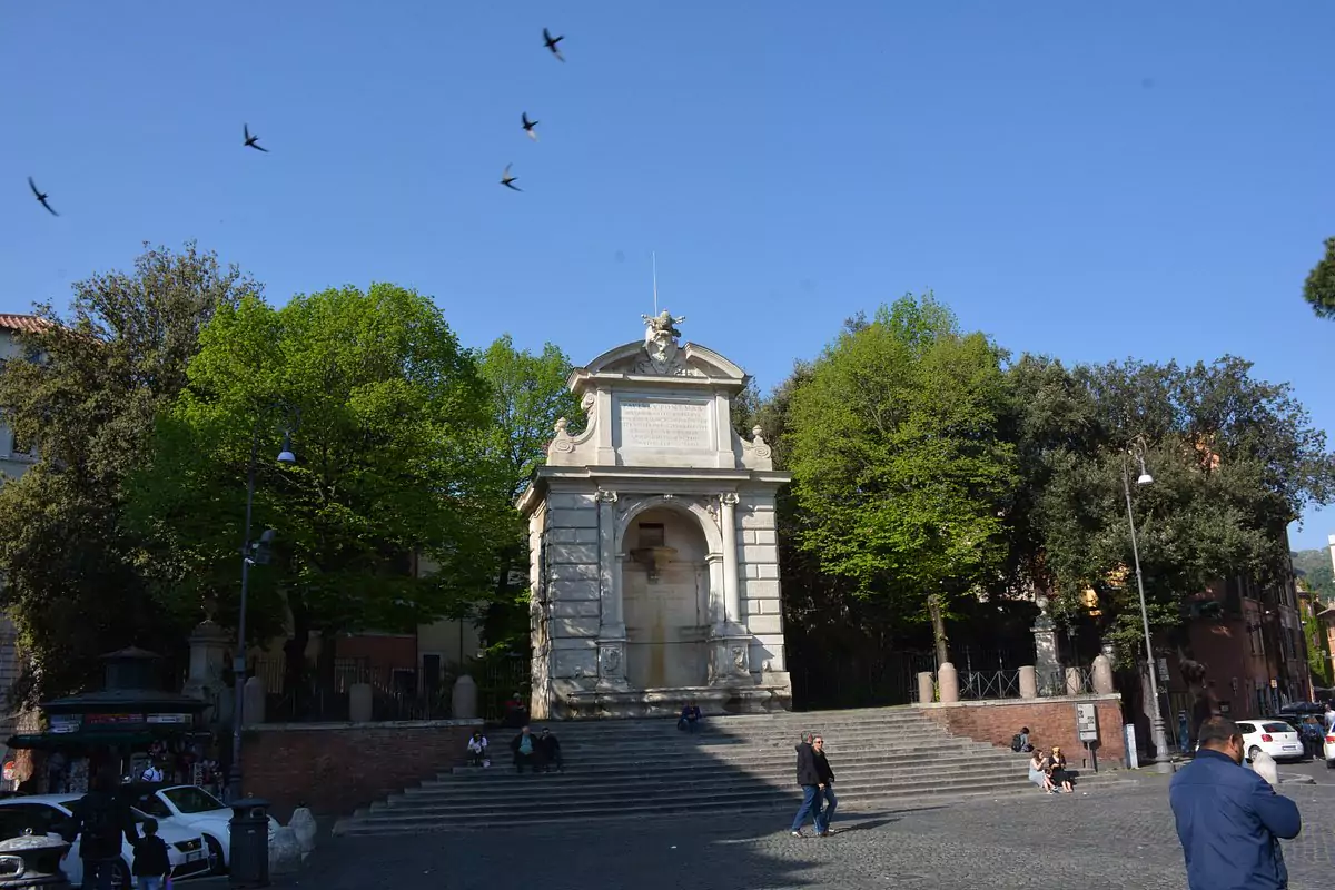 The ornate Fountain of Ponte Sisto in Rome, featuring carved stone sculptures and water flowing into a basin, set against the backdrop of the historic Ponte Sisto bridge. The classical design highlights the city’s rich artistic heritage.