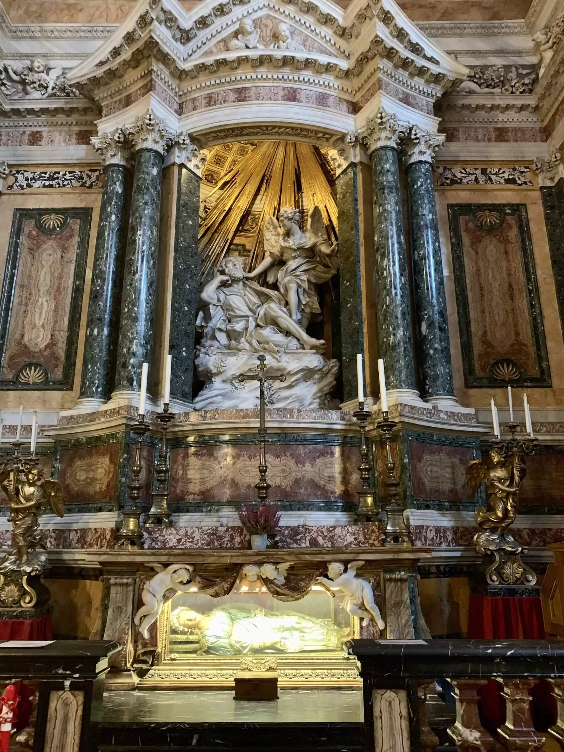 The Ecstasy of Saint Teresa sculpture by Bernini, viewed inside Santa Maria della Vittoria in Rome, showing the dramatic marble figures bathed in golden light from hidden rays, set within an ornately decorated chapel.