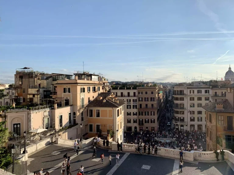 Panoramic view of Rome from the top of the Spanish Steps, featuring historic rooftops, church domes, and winding streets stretching out under a bright, sunny sky.