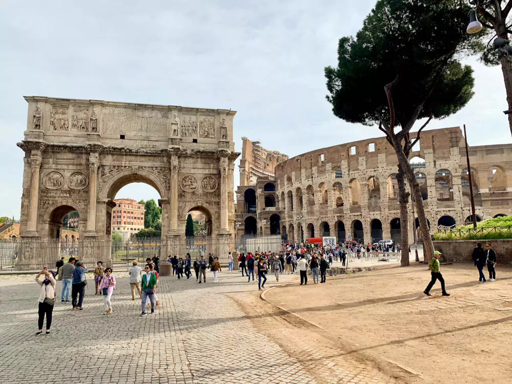 The Colosseum and Arch of Constantine in Rome, Italy, standing side by side with the ancient amphitheater’s massive stone arches and the triumphal arch’s intricate carvings framed under a clear blue sky.